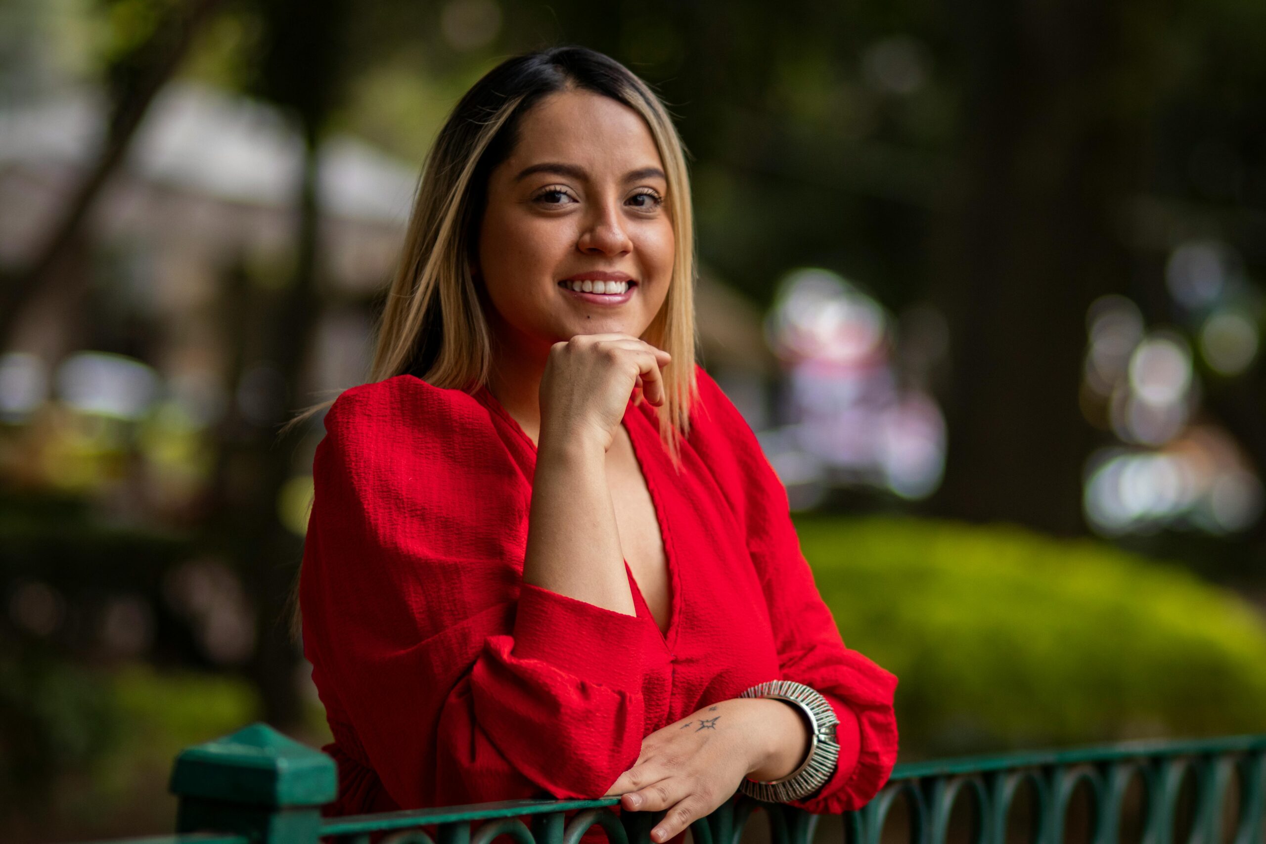 Young woman in red blouse, smiling outdoors against a blurred background.